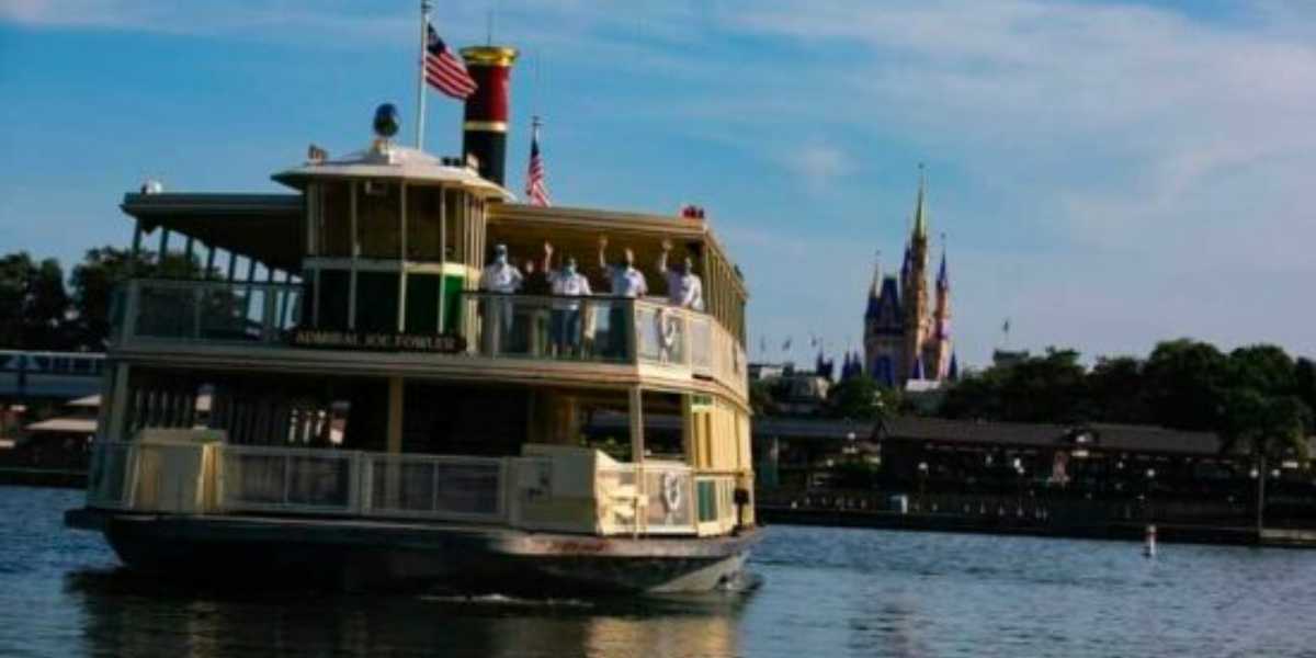 A ferryboat named "Admiral Joe Fowler" crosses a lake with passengers on board, American flags flying, and Cinderella Castle visible in the background under a blue sky.