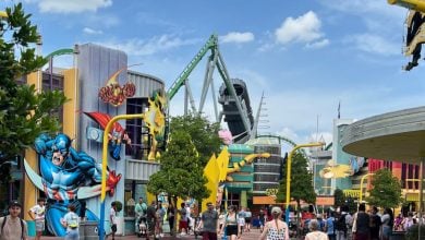People walk along a colorful Islands of Adventure theme park street with comic book superhero decorations, including Captain America and the Hulk. A roller coaster track is visible in the background under a partly cloudy blue sky.