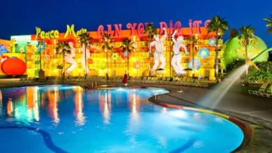 A brightly lit, retro-themed Pop Century hotel with colorful 1960s decor—large peace signs, flowers, and dancing figures—is reflected in a glowing outdoor pool at dusk. Palm trees line the vibrant scene.