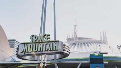 The image shows the Space Mountain attraction sign with stylized green and white letters, in front of the iconic white, futuristic building of the ride at Disneyland, under a clear sky.