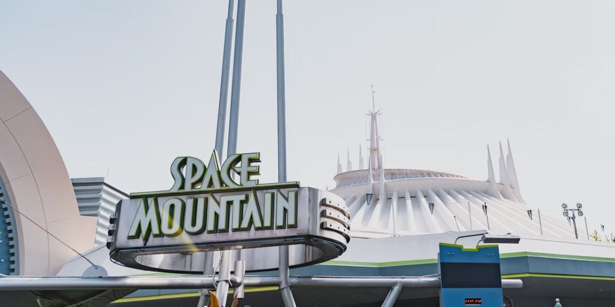 The image shows the Space Mountain attraction sign with stylized green and white letters, in front of the iconic white, futuristic building of the ride at Disneyland, under a clear sky.