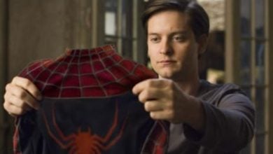 A young man with short brown hair holds up a red and blue Spider-Man suit, looking at it thoughtfully while standing indoors.