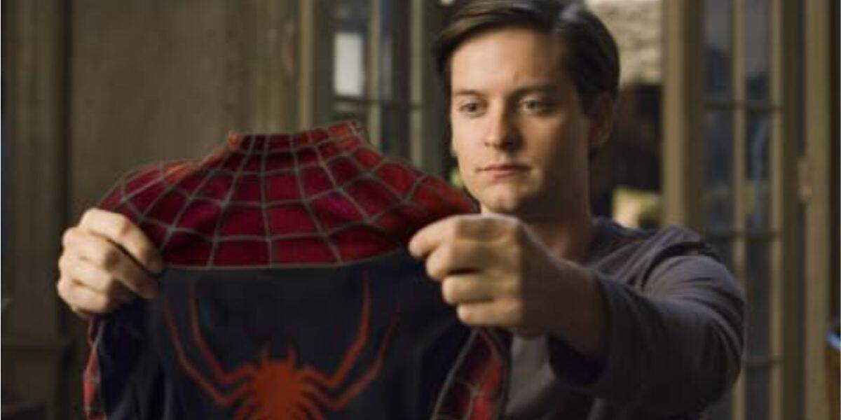 A young man with short brown hair holds up a red and blue Spider-Man suit, looking at it thoughtfully while standing indoors.