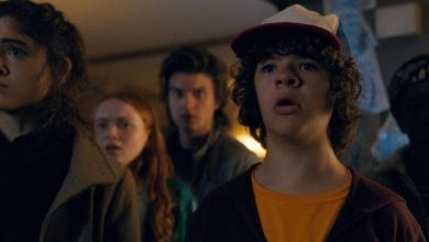 Five teenagers stand close together indoors, looking ahead with expressions of shock and concern. The lighting is dim and the mood appears tense.