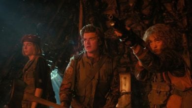 Three people in rugged clothing stand alert in a dimly lit, stranger things-inspired setting. One woman aims a rifle, another wields an axe, and the man in the center looks intensely ahead as eerie light filters through gaps behind them.