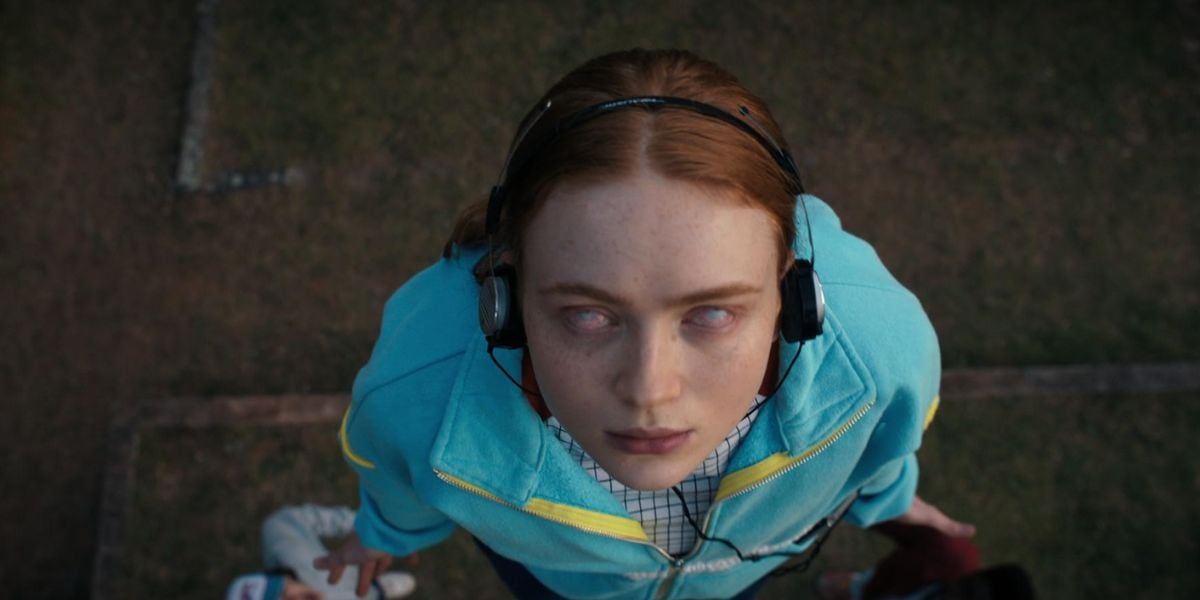 A girl with red hair and pale skin wears headphones and a blue jacket, looking upward with blank, cloudy eyes, standing on grass. The image is shot from above, giving a dramatic and intense perspective.