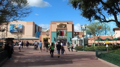 Crowds of people walk toward the entrance of Disney’s Hollywood Studios, with the "Star Wars Launch Bay" sign visible above the doors. Greenery and lampposts line the walkway under a blue sky with scattered clouds.