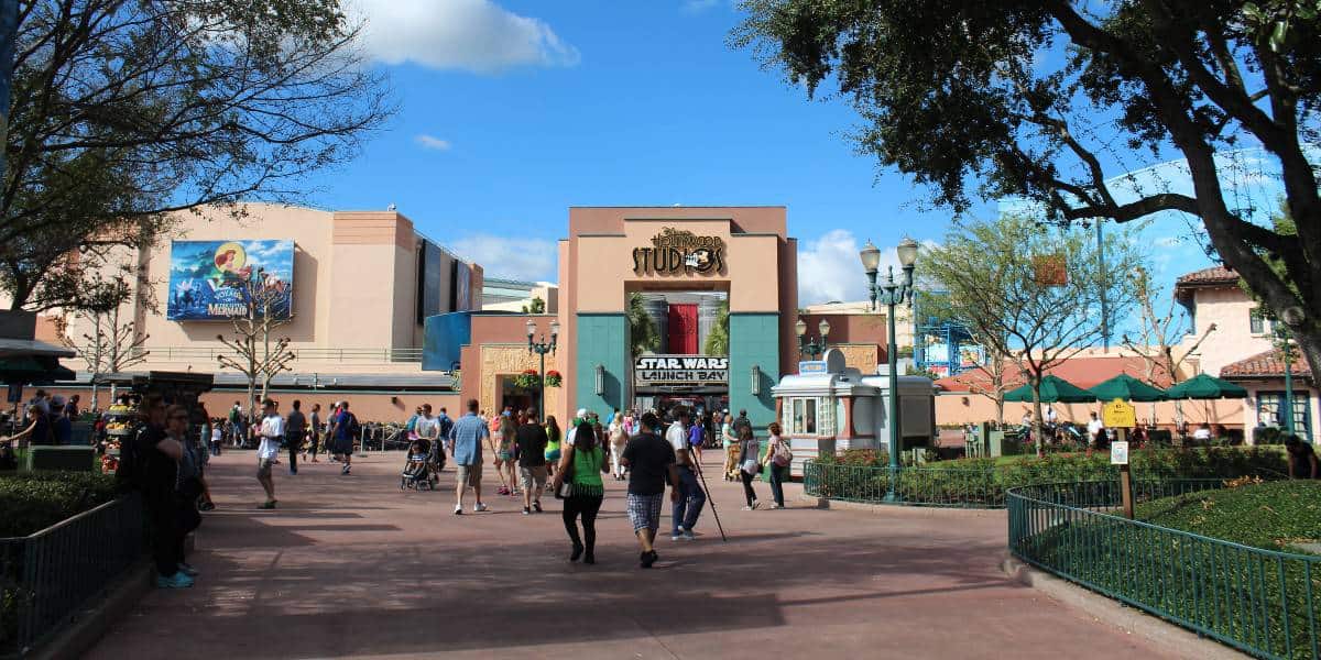 Crowds of people walk toward the entrance of Disney’s Hollywood Studios, with the "Star Wars Launch Bay" sign visible above the doors. Greenery and lampposts line the walkway under a blue sky with scattered clouds.