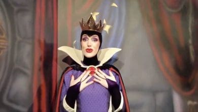A person dressed as the Evil Queen from Snow White, wearing a crown, purple and black robe, and dramatic makeup, poses with hands clasped near a red gem on their chest, in front of a blurred backdrop.