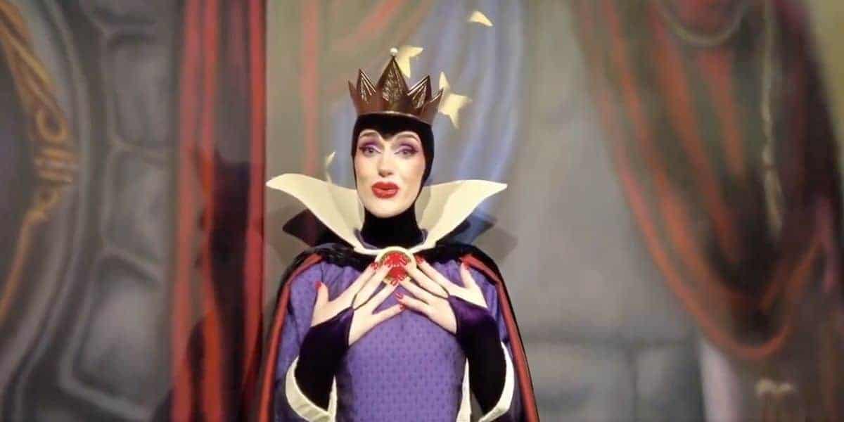 A person dressed as the Evil Queen from Snow White, wearing a crown, purple and black robe, and dramatic makeup, poses with hands clasped near a red gem on their chest, in front of a blurred backdrop.