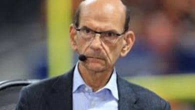 A serious-looking man with glasses, wearing a headset microphone, blue shirt, and dark jacket, sits indoors with a blurred background.
