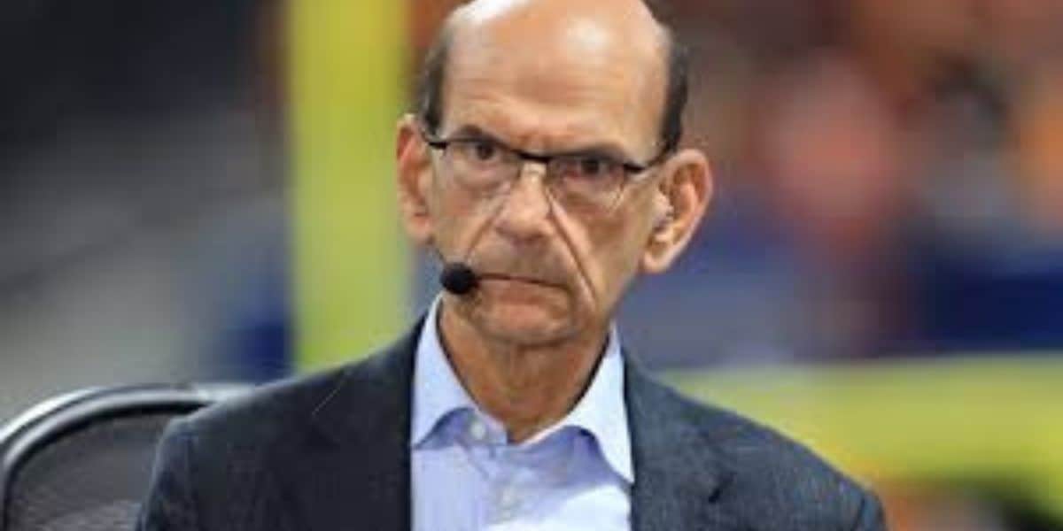 A serious-looking man with glasses, wearing a headset microphone, blue shirt, and dark jacket, sits indoors with a blurred background.