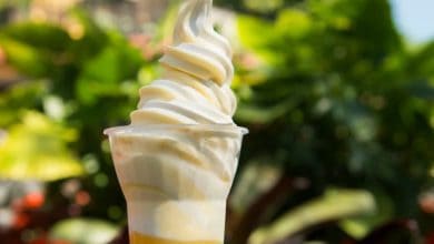 A close-up of a vanilla soft-serve ice cream swirl in a clear plastic cup, inspired by classic Disney treats, set against a blurred green leafy background.