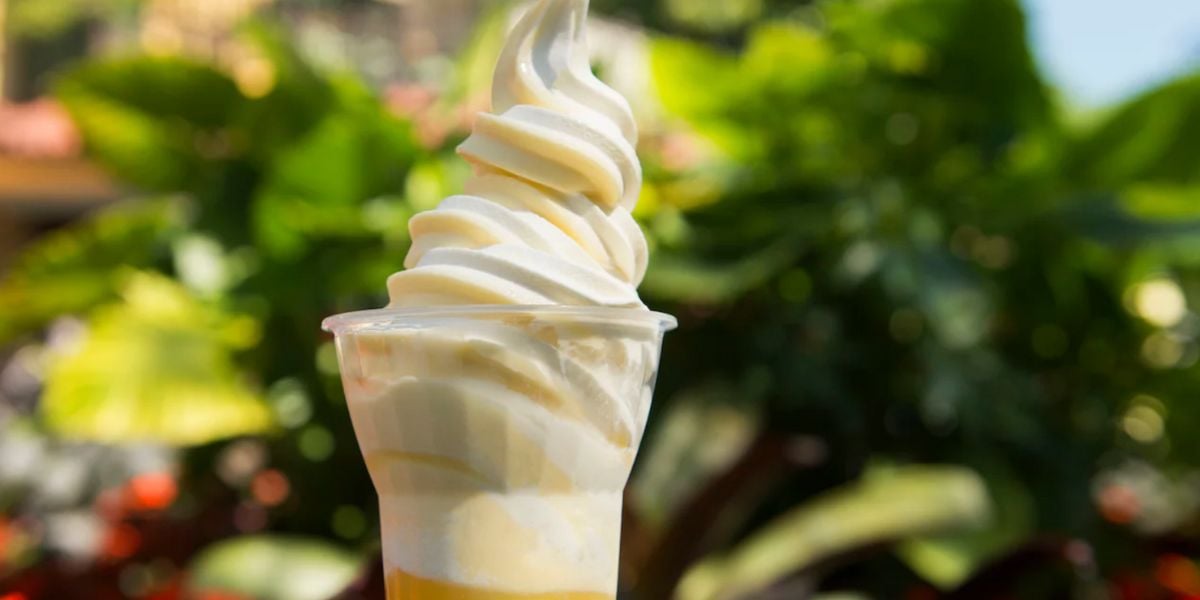 A close-up of a vanilla soft-serve ice cream swirl in a clear plastic cup, inspired by classic Disney treats, set against a blurred green leafy background.
