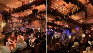 A dimly lit, medieval-themed restaurant filled with people dining at wooden tables. The decor features candles, cobwebs, stone walls, and shelves displaying eerie objects and glowing lights.