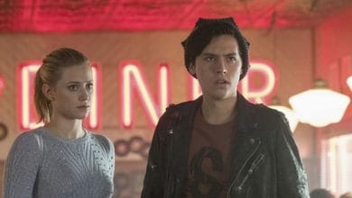 Two young people stand inside a retro diner with neon red "DINER" sign in the background; the woman has blonde hair in a ponytail and wears a light sweater, the man wears a dark jacket and beanie, both look concerned.