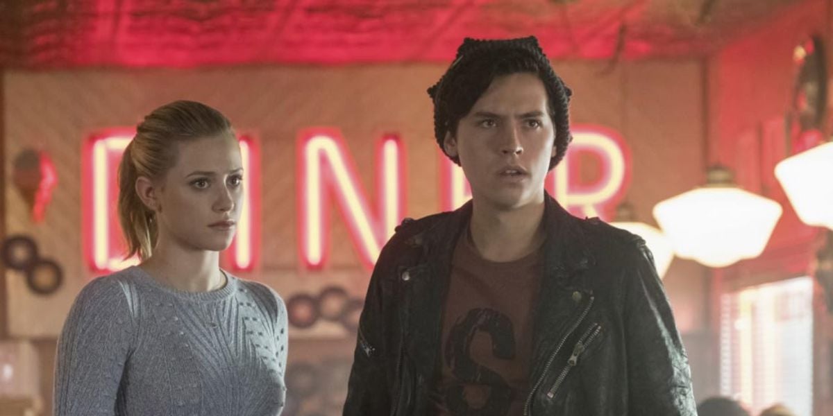 Two young people stand inside a retro diner with neon red "DINER" sign in the background; the woman has blonde hair in a ponytail and wears a light sweater, the man wears a dark jacket and beanie, both look concerned.