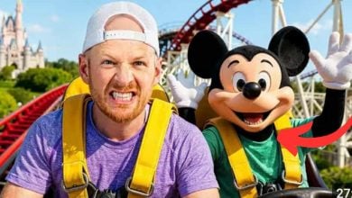 Dude perfect and an AI person in a Mickey Mouse costume ride a roller coaster together, both wearing yellow harnesses. The man looks nervous while Mickey Mouse appears excited with raised hands. A castle is visible in the background.