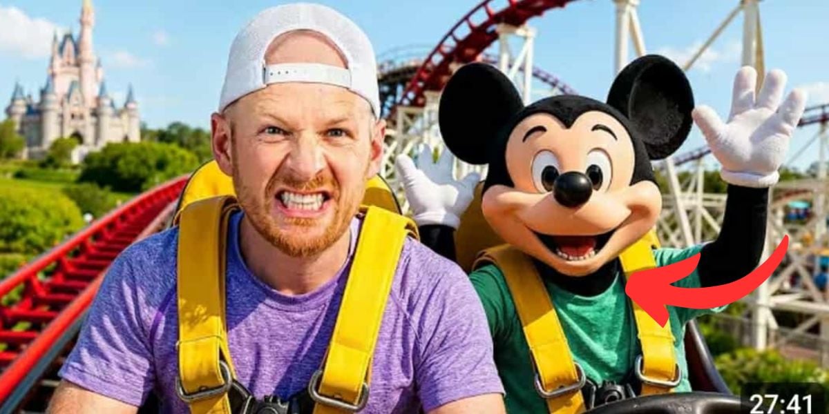 Dude perfect and an AI person in a Mickey Mouse costume ride a roller coaster together, both wearing yellow harnesses. The man looks nervous while Mickey Mouse appears excited with raised hands. A castle is visible in the background.