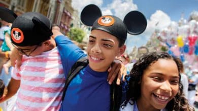 Three smiling children, two wearing Disney Mickey Mouse ears, walk arm-in-arm outdoors on a sunny day in an amusement park. Colorful balloons and other people are visible in the background.