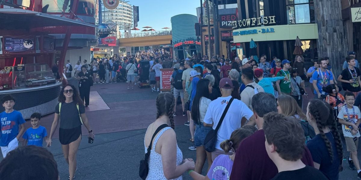 A large crowd of people waits in a long line outside various restaurants and shops at an outdoor entertainment complex on a sunny day.