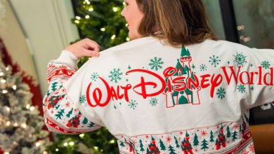 A woman faces away, showing a festive holiday sweater in white with red and green patterns and "Walt Disney World" across the back, standing near a decorated Christmas tree.