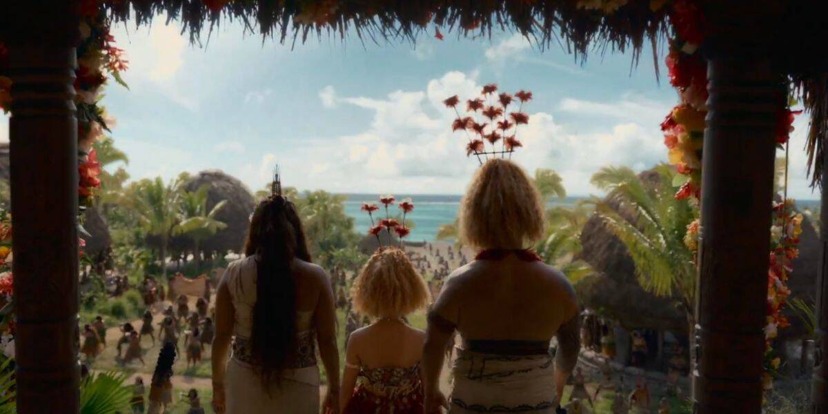 Three people in traditional Polynesian attire stand under a thatched roof, facing a tropical village with palm trees and huts by the ocean, as a crowd gathers in the sunny background, evoking a scene from Live Action Moana.