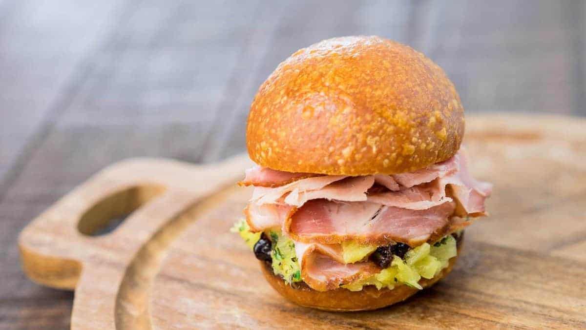 A ham sandwich with shredded ham, pineapple, and raisins on a round, golden bun sits on a wooden cutting board.