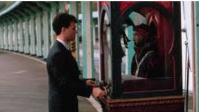 A man in a suit stands in front of a vintage fortune teller machine on an empty, covered boardwalk, inserting a coin or interacting with the machine.