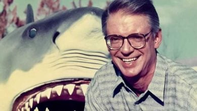 A smiling man wearing glasses and a patterned shirt stands in front of a large, open-mouthed shark model or statue, with trees blurred in the background.