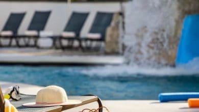 A poolside scene with a lounge chair holding a sun hat and sunglasses in the foreground, pool noodles on the ground, and several empty lounge chairs near a pool with water cascading in the background.