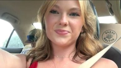 A young woman with blonde hair and a red top smiles while taking a selfie inside a car. The car seat cover behind her has a circular logo with text that is partially visible. Sunlight comes through the windows.