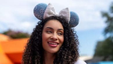 A woman with curly hair smiles while wearing large blue mouse ears with a white bow at Epcot. The background is outdoors, featuring blurred trees and bright orange shapes.