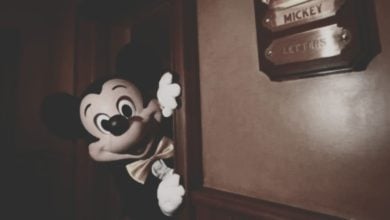 A person in a Mickey Mouse costume peeks playfully around a wooden doorway. A brass plaque on the wall nearby reads "Mickey" and "Letters." The scene is warmly lit and has a nostalgic feel.