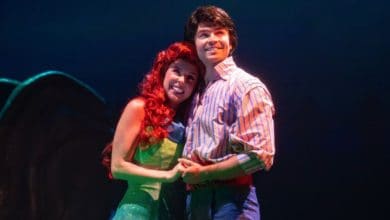 Two actors dressed as Ariel and Prince Eric from Disney's "The Little Mermaid" musical stand close together on stage, smiling and holding hands under dramatic blue lighting.