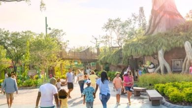 A diverse group of people, including families and children, walk along a sunlit path surrounded by greenery and themed structures in a lively outdoor amusement or theme park.