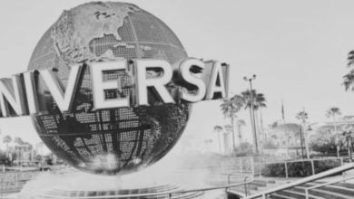 A large globe sculpture with the word "UNIVERSAL" in bold letters stands at the entrance of Universal Studios, surrounded by palm trees and mist, in a black-and-white photo.