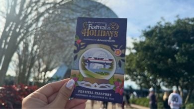 A Holidays Passport is held up in front of Spaceship Earth, capturing the festive fun of EPCOT on a bright, sunny day.