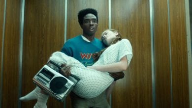 Two teens reenact an '80s movie scene, one in a hospital gown and one with a boombox, inside a retro elevator.