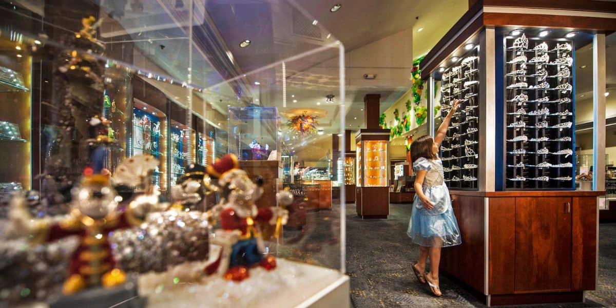 A little Disney fan in a blue dress picks out sunglasses inside a bright Disney Springs shop filled with glittering collectibles.