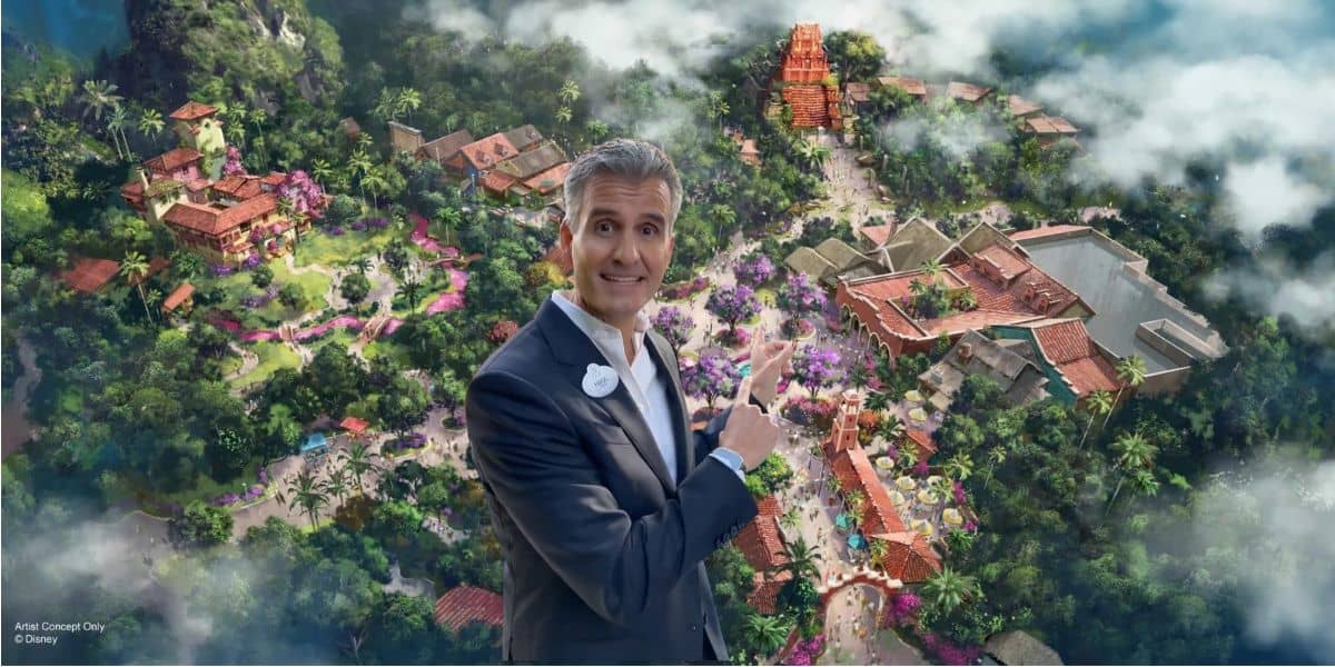 Josh D’Amaro stands smiling and gesturing before lush Disney Parks concept art, inspiring excitement for future attractions.