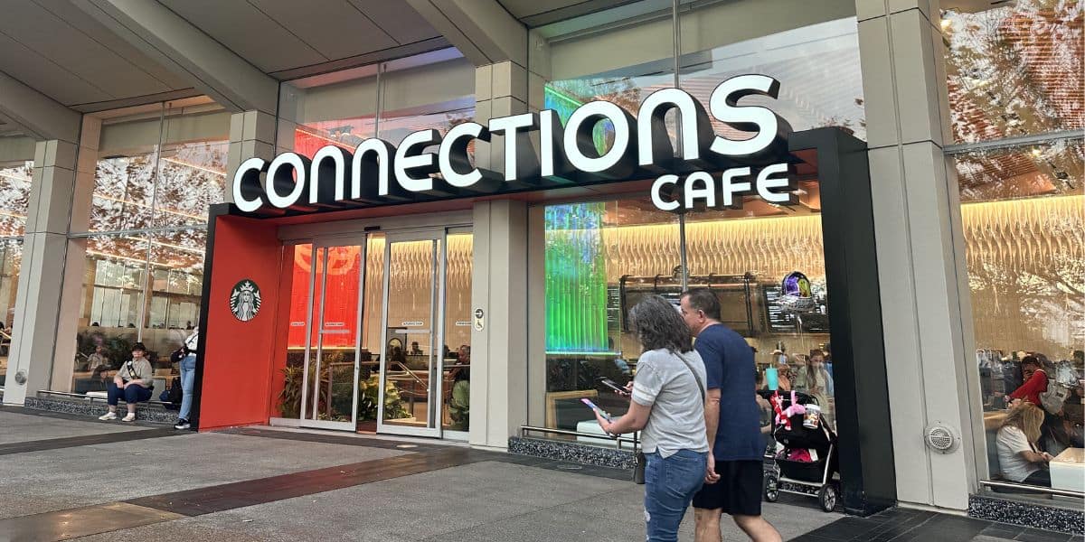 Guests stroll past the glass doors of Connections Café at EPCOT, featuring a Starbucks logo and plenty of inviting seating inside.