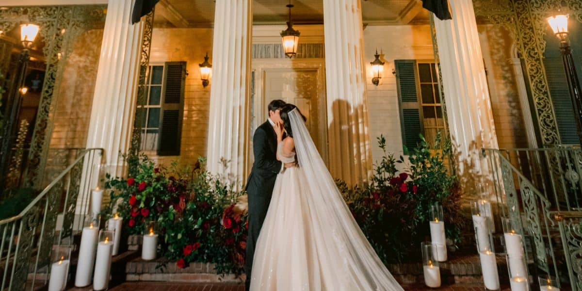 A newlywed couple kisses under dazzling lights outside a grand, castle-like venue reminiscent of Disney’s magical parks at night.
