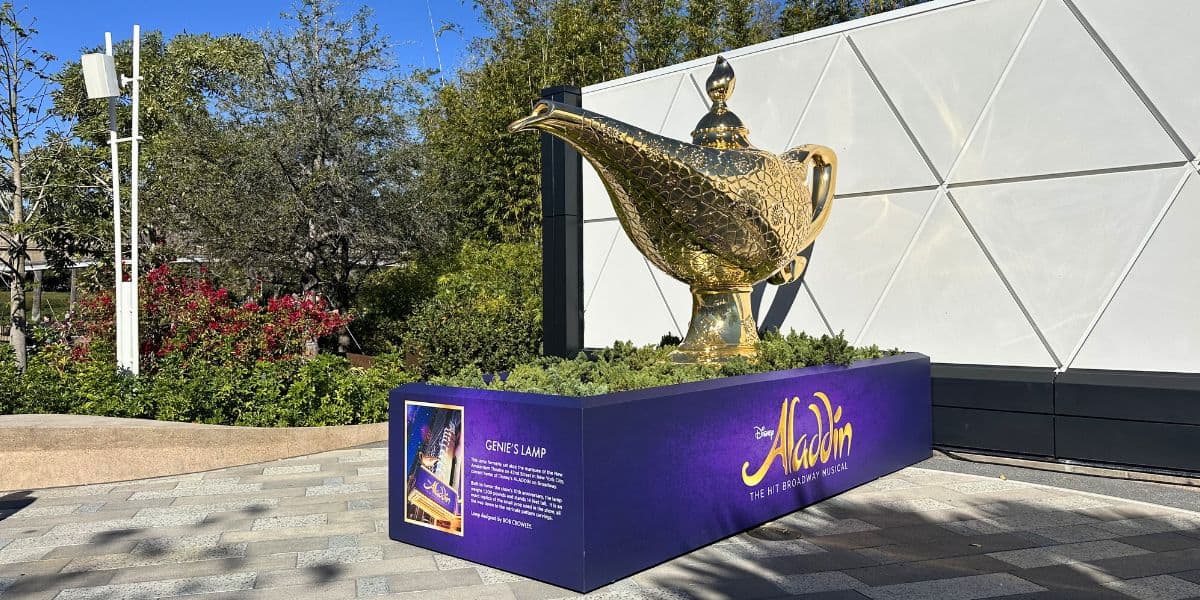 Golden Genie Lamp centerpiece gleams atop a vibrant Aladdin display in Disney Park, framed by lush trees and a white backdrop.