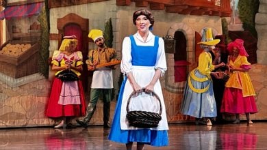 Belle in her signature blue dress holds a basket on stage, surrounded by villagers and a lively marketplace from Beauty and the Beast.