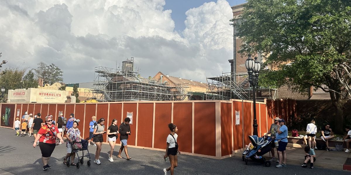Guests stroll by a construction zone bordered by scaffolding and a tall brown fence inside Disney Park on a sunny day.