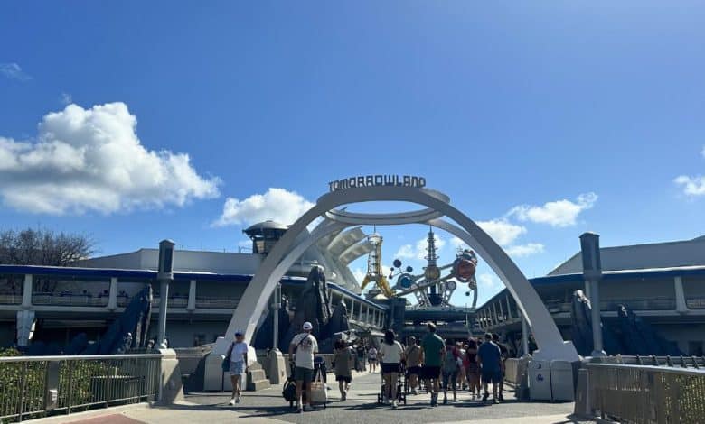 Guests stroll beneath the iconic Tomorrowland arch at Disneyland, under sunny blue skies, passing an area hinting at future updates.