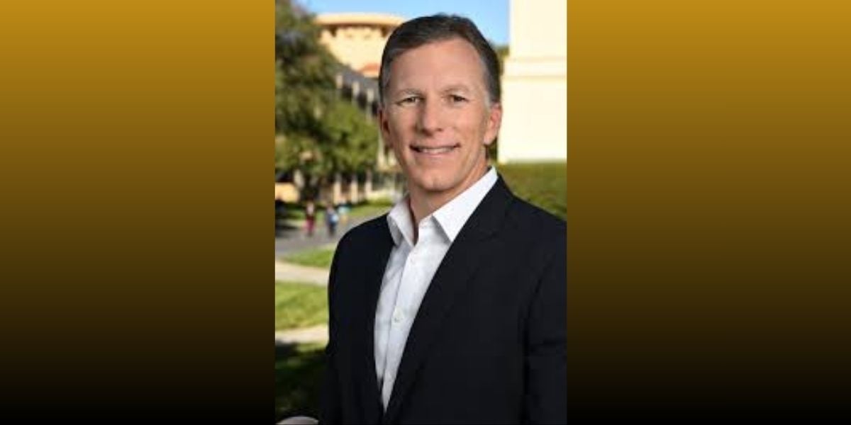 A man in a dark suit jacket and white shirt grins along a Main Street-style avenue, with whimsical Disney-like buildings behind him.