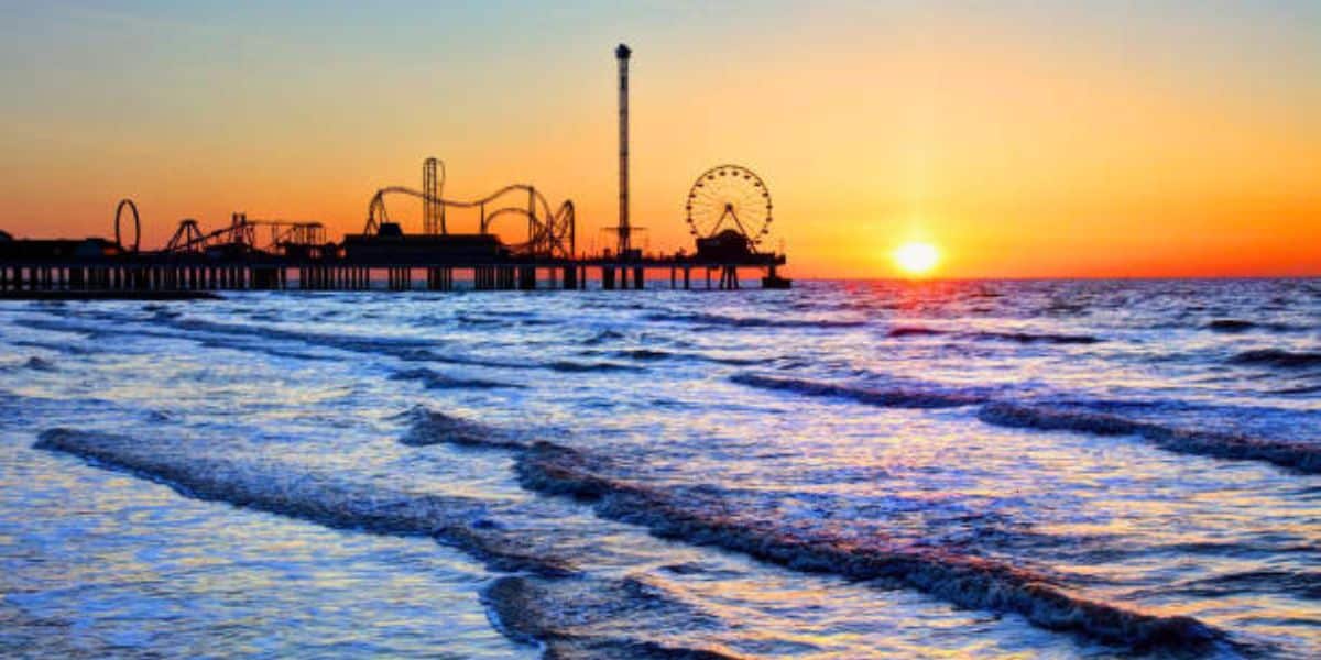 Galveston Island Historic Pleasure Pier at sunset, with thrill rides and the roller coaster silhouetted over crashing ocean waves.