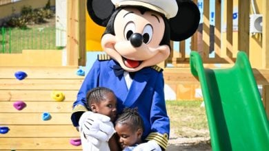 Mickey Mouse shares a warm hug with two kids beside a playground slide under the sunny skies of Disney World.